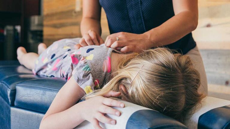 A chiropractor uses hands to gently adjust a person's back on a cushioned table in a well-lit clinical setting.